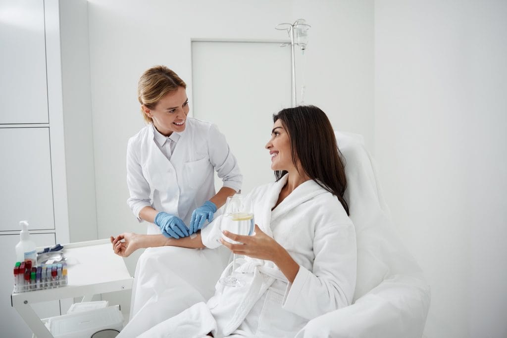 Smiling doctor attaching intravenous drip on lady hand, Portrait of charming woman sitting in armchair and holding glass of lemon water while doctor in sterile gloves checking IV infusion | Citidrips in Wilmington, DE